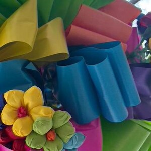 Op de afbeelding: Een kleurrijke krans gemaakt van meerdere linten in tinten groen, geel, roze, blauw en paars. De krans is versierd met kleine, handgemaakte papieren bloemen in geel, groen en roze.