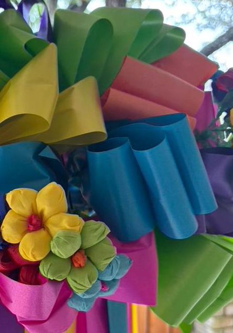 Op de afbeelding: Een kleurrijke krans gemaakt van meerdere linten in tinten groen, geel, roze, blauw en paars. De krans is versierd met kleine, handgemaakte papieren bloemen in geel, groen en roze.