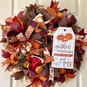 May include: A decorative wreath featuring autumn-colored mesh ribbons in shades of brown, orange, and burgundy. It includes fabric pie slice ornaments and a rectangular sign with two pumpkins and the text, gather TOGETHER GIVE THANKS and eat PIE.