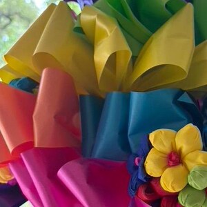 Op de afbeelding: Een kleurrijke regenboog lintdecoratie met gele, groene, blauwe, oranje, roze en paarse linten. De linten zijn samen gebonden in een grote strik met een klein geel bloemetje in het midden.