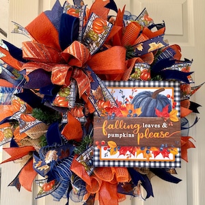 May include: A decorative wreath featuring orange and navy blue mesh ribbons, patterned fabric ribbons with pumpkin and leaf prints, and a central rectangular sign that reads falling leaves & pumpkins please with a blue pumpkin illustration.