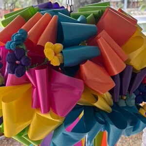 Op de afbeelding: Een kleurrijke regenboog-decoratie gemaakt van meerdere lagen lint, met een cluster van papieren bloemen in het midden.