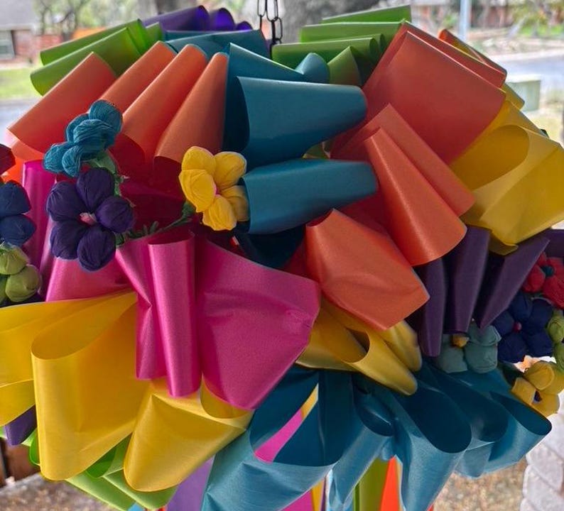 Op de afbeelding: Een kleurrijke regenboog-decoratie gemaakt van meerdere lagen lint, met een cluster van papieren bloemen in het midden.
