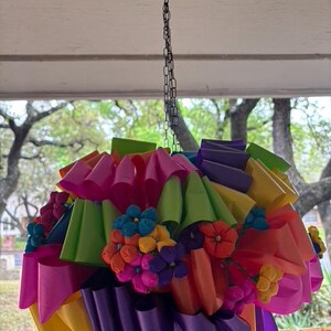 Op de afbeelding: Een kleurrijke papieren pi&ntilde;ata met een ketting die aan het plafond hangt. De pi&ntilde;ata is gemaakt van meerdere lagen cr&ecirc;pepapier in verschillende kleuren, waaronder roze, groen, paars, geel en oranje. De pi&ntilde;ata is versierd met kleine papieren bloemen in tinten roze, blauw, geel en paars.
