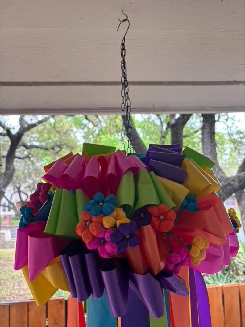 Op de afbeelding: Een kleurrijke papieren pi&ntilde;ata met een ketting die aan het plafond hangt. De pi&ntilde;ata is gemaakt van meerdere lagen cr&ecirc;pepapier in verschillende kleuren, waaronder roze, groen, paars, geel en oranje. De pi&ntilde;ata is versierd met kleine papieren bloemen in tinten roze, blauw, geel en paars.