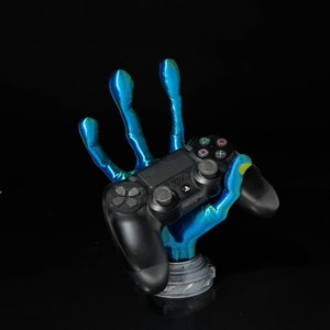 Könnte beinhalten: Eine blaue und grüne 3D-gedruckte Hand hält einen schwarzen PlayStation 4-Controller. Die Hand ist mit drei ausgestreckten Fingern und Daumen und kleinem Finger gekrümmt.