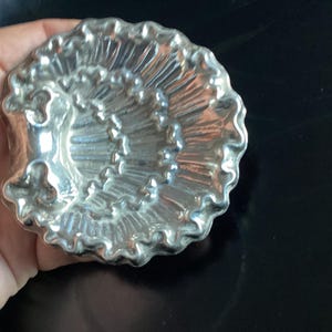 Puede incluir: Un molde plateado con forma de concha marina con un dise&ntilde;o detallado y acanalado. El molde tiene un borde festoneado y se sostiene en una mano sobre un fondo oscuro. Este art&iacute;culo decorativo probablemente se utiliza para hornear o hacer manualidades.