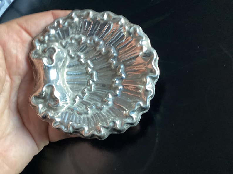 Puede incluir: Un molde plateado con forma de concha marina con un dise&ntilde;o detallado y acanalado. El molde tiene un borde festoneado y se sostiene en una mano sobre un fondo oscuro. Este art&iacute;culo decorativo probablemente se utiliza para hornear o hacer manualidades.