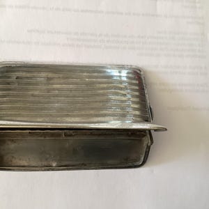 May include: A rectangular, silver-toned metal object with a ridged, translucent top and a lower compartment. The object has a small, curved lip extending from the bottom. The item is likely a vintage or antique automotive part.