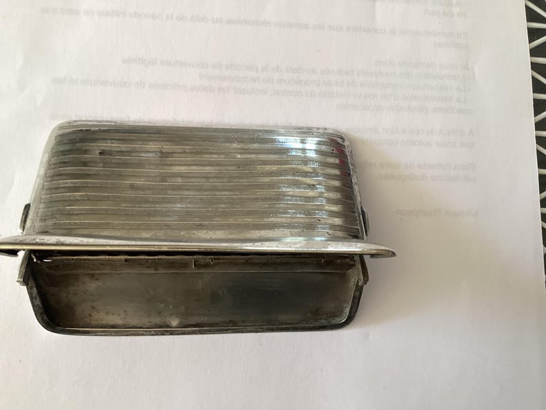 May include: A rectangular, silver-toned metal object with a ridged, translucent top and a lower compartment. The object has a small, curved lip extending from the bottom. The item is likely a vintage or antique automotive part.