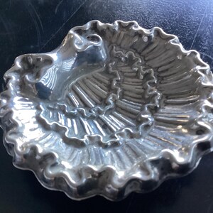 Puede incluir: Un plato plateado en forma de concha con un dise&ntilde;o en espiral. El plato tiene un borde con volantes y una peque&ntilde;a zona elevada en un lado. El plato es probablemente de metal y est&aacute; sobre una superficie oscura.