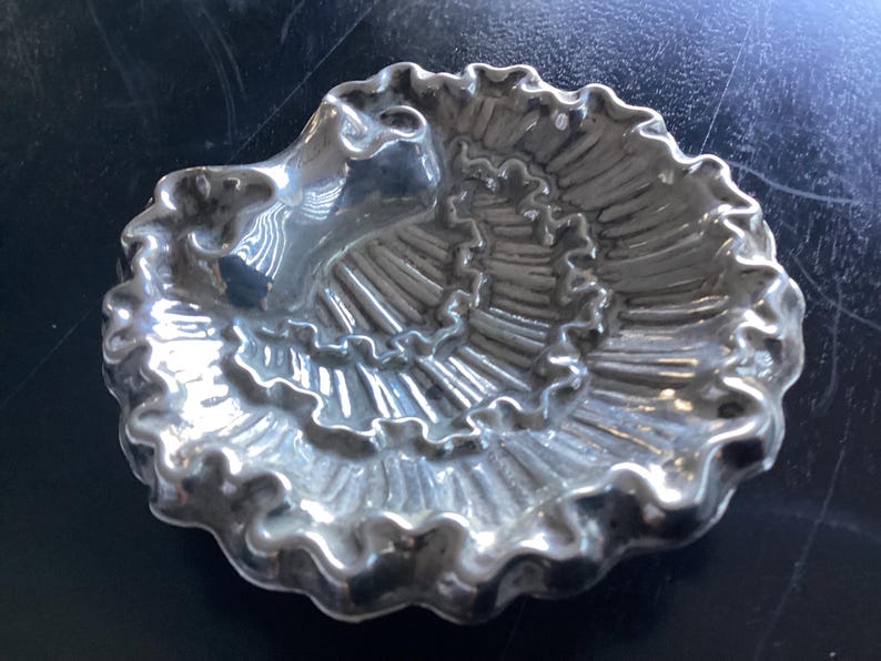 Puede incluir: Un plato plateado en forma de concha con un dise&ntilde;o en espiral. El plato tiene un borde con volantes y una peque&ntilde;a zona elevada en un lado. El plato es probablemente de metal y est&aacute; sobre una superficie oscura.