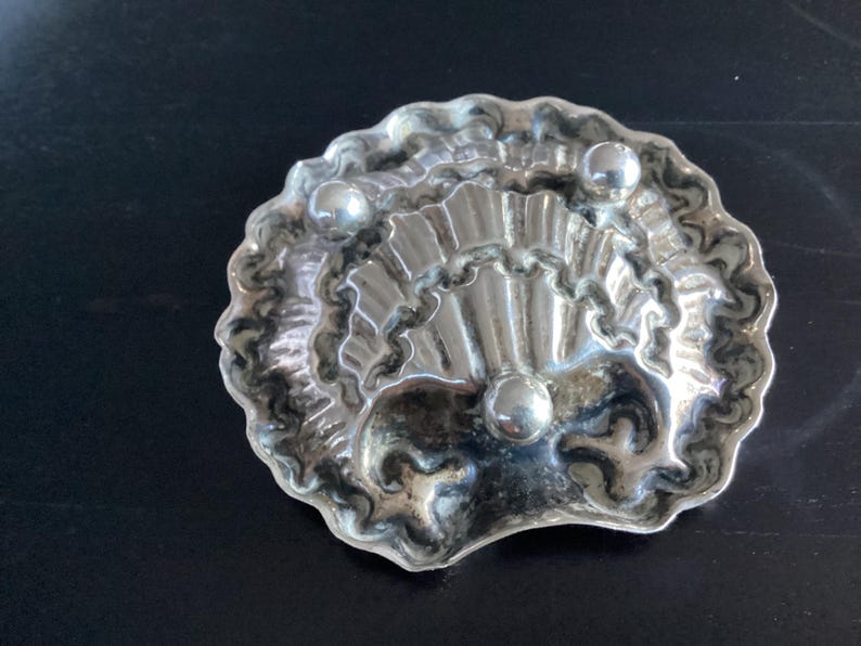 Puede incluir: Un plato plateado en forma de concha con tres peque&ntilde;os acentos redondos plateados. El plato tiene un borde ondulado y una superficie texturizada detallada. El dise&ntilde;o recuerda a una concha de vieira.