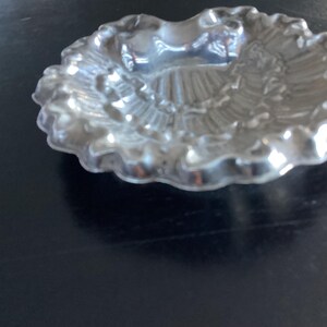 Puede incluir: Un peque&ntilde;o plato plateado con borde festoneado. El plato tiene un dise&ntilde;o detallado y texturizado en el interior, con una depresi&oacute;n central. El plato es probablemente de metal y se encuentra sobre una superficie oscura.