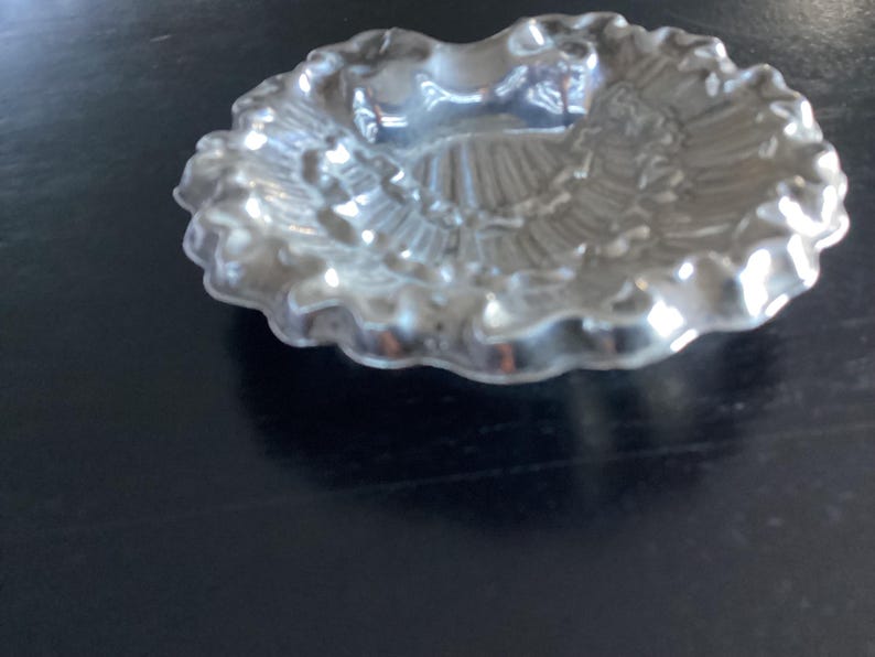 Puede incluir: Un peque&ntilde;o plato plateado con borde festoneado. El plato tiene un dise&ntilde;o detallado y texturizado en el interior, con una depresi&oacute;n central. El plato es probablemente de metal y se encuentra sobre una superficie oscura.