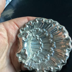 Puede incluir: Objeto decorativo plateado en forma de concha con un dise&ntilde;o detallado y acanalado. El objeto tiene un patr&oacute;n en espiral y un borde con volantes. El art&iacute;culo se sostiene en una mano sobre un fondo oscuro.