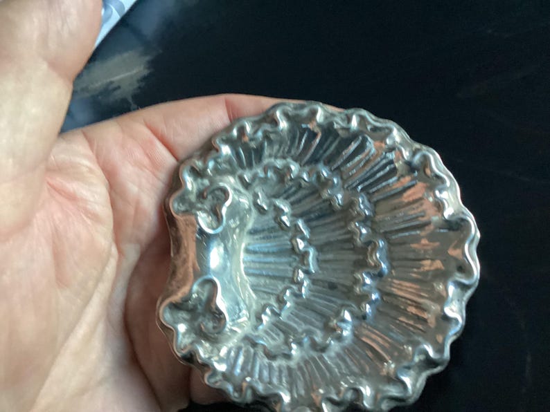 Puede incluir: Objeto decorativo plateado en forma de concha con un dise&ntilde;o detallado y acanalado. El objeto tiene un patr&oacute;n en espiral y un borde con volantes. El art&iacute;culo se sostiene en una mano sobre un fondo oscuro.