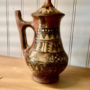 Può includere: Una brocca in terracotta con coperchio abbinato e manico laterale, caratterizzata da un disegno dipinto a mano in nero e crema. La superficie è decorata con motivi geometrici, tra cui triangoli, linee e quadrati a scacchiera.