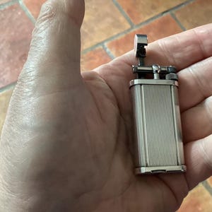 May include: A silver rectangular lighter with vertical grooved detailing. The lighter features a hinged top and a small ignition wheel. The lighter is held in an open hand, highlighting its design and size.