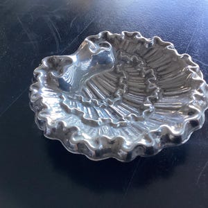 Pode incluir: Um prato prateado em forma de concha com uma borda ondulada. O prato tem uma depressão central e um design texturizado e canelado que irradia para fora. O prato é provavelmente feito de metal e está sobre uma superfície escura.