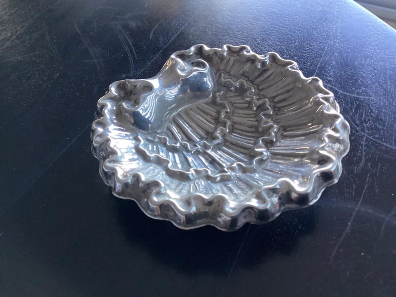 Puede incluir: Un plato plateado en forma de concha con un borde con volantes. El plato tiene una depresi&oacute;n central y un dise&ntilde;o texturizado y acanalado que irradia hacia afuera. El plato probablemente est&aacute; hecho de metal y est&aacute; sobre una superficie oscura.