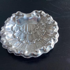 Puede incluir: Un peque&ntilde;o plato plateado con borde festoneado. El plato tiene un interior texturizado y acanalado, y la forma general se asemeja a una concha. El plato es probablemente de metal y refleja la luz.
