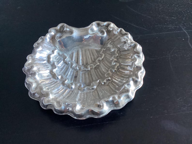 Puede incluir: Un peque&ntilde;o plato plateado con borde festoneado. El plato tiene un interior texturizado y acanalado, y la forma general se asemeja a una concha. El plato es probablemente de metal y refleja la luz.