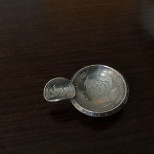 Può includere: Una moneta d'argento a forma di piccola ciotola. La moneta presenta il ritratto di un uomo con il testo "LIG ONE" attorno al bordo.