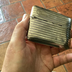 May include: A rectangular, silver-toned metal case with horizontal striped detailing. The case has a hinged lid and an oval accent on the top. The item is held in a hand, suggesting its size and portability. The case appears to be vintage.
