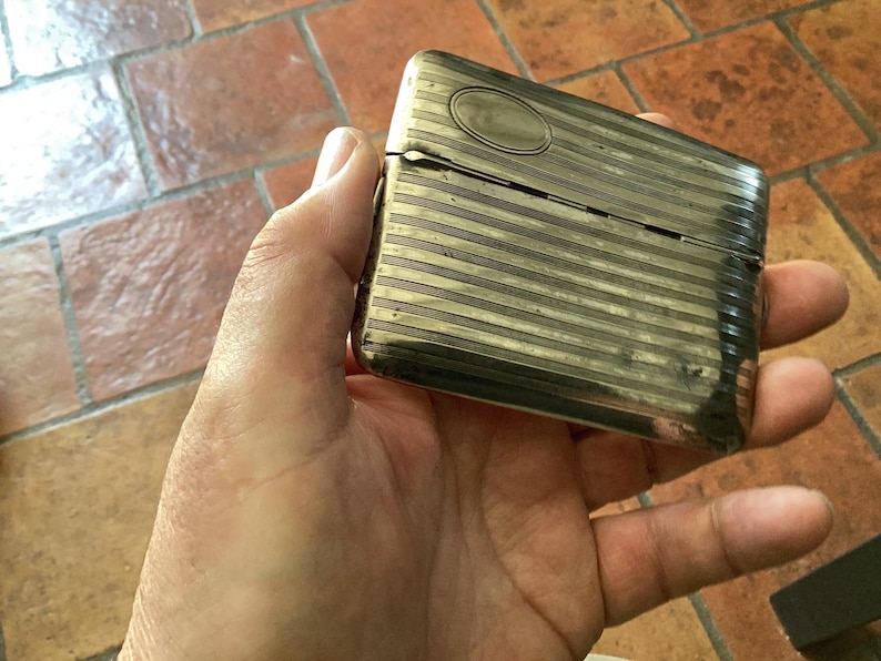 May include: A rectangular, silver-toned metal case with horizontal striped detailing. The case has a hinged lid and an oval accent on the top. The item is held in a hand, suggesting its size and portability. The case appears to be vintage.
