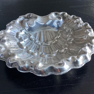 Puede incluir: Un peque&ntilde;o plato decorativo de color plateado con borde festoneado. El plato tiene un interior texturizado y acanalado y una forma poco profunda y redondeada. El plato es probablemente de metal y se encuentra sobre una superficie oscura.
