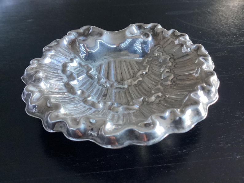 Puede incluir: Un peque&ntilde;o plato decorativo de color plateado con borde festoneado. El plato tiene un interior texturizado y acanalado y una forma poco profunda y redondeada. El plato es probablemente de metal y se encuentra sobre una superficie oscura.