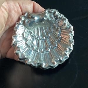 Puede incluir: Un plato plateado en forma de concha con un borde con volantes. El plato tiene una superficie detallada y texturizada y se sostiene en una mano. El plato probablemente se usa para servir art&iacute;culos peque&ntilde;os o como pieza decorativa.