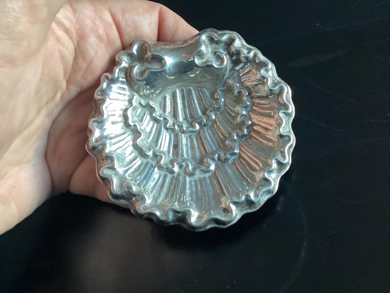 Puede incluir: Un plato plateado en forma de concha con un borde con volantes. El plato tiene una superficie detallada y texturizada y se sostiene en una mano. El plato probablemente se usa para servir art&iacute;culos peque&ntilde;os o como pieza decorativa.