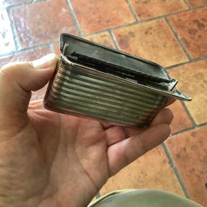 May include: A small, rectangular silver-toned box with a hinged lid, held open. The box has horizontal striped detailing and a clasp. The background is a tiled surface.