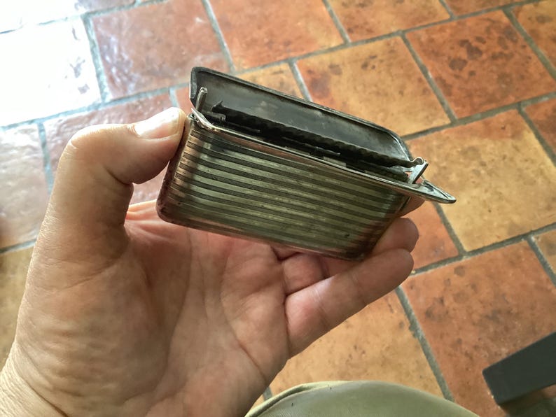 May include: A small, rectangular silver-toned box with a hinged lid, held open. The box has horizontal striped detailing and a clasp. The background is a tiled surface.