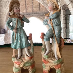May include: Two porcelain figurines of children in pastel blue and gold outfits, each holding a staff. The figures stand on ornate, colorful bases with leaf and floral designs. Both wear straw hats.