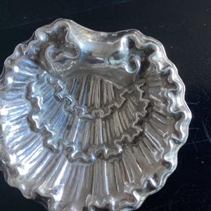 Puede incluir: Un plato plateado en forma de concha con un dise&ntilde;o detallado y acanalado. El plato tiene un borde festoneado y una depresi&oacute;n central. La forma general recuerda a una concha, con una superficie texturizada y un brillo reflectante.