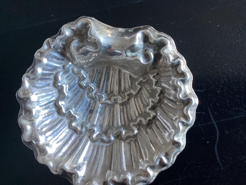 Puede incluir: Un plato plateado en forma de concha con un dise&ntilde;o detallado y acanalado. El plato tiene un borde festoneado y una depresi&oacute;n central. La forma general recuerda a una concha, con una superficie texturizada y un brillo reflectante.