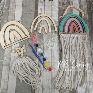 May include: A DIY rainbow wall hanging kit. The kit includes unfinished wooden rainbow shapes, paint pots, paintbrushes, wooden beads, and macrame cord. One completed rainbow wall hanging is shown with a turquoise, purple, and coral color scheme.