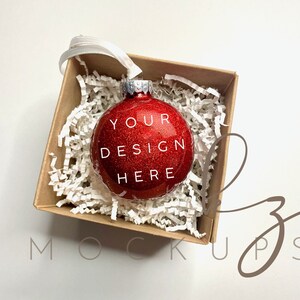 Könnte beinhalten: Ein roter Glitzer-Weihnachtsbaumschmuck mit dem Text "YOUR DESIGN HERE" in einer weißen Box gefüllt mit weißem zerknülltem Papier.