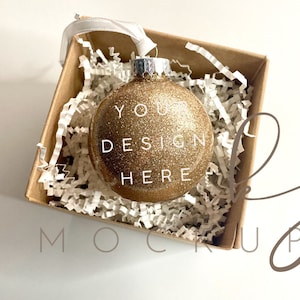 May include: A gold glitter Christmas ornament with the text "YOUR DESIGN HERE" in a white box filled with white shredded paper.