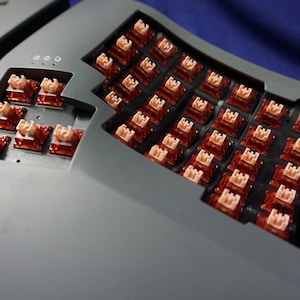 Puede incluir: Primer plano de un teclado gris con un diseño curvo. El teclado tiene una variedad de teclas, incluyendo un teclado numérico y una fila de funciones. Las teclas son todas negras y tienen un acabado mate. El teclado está hecho de un plástico duradero y tiene un diseño elegante y moderno.