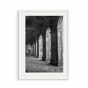 Op de afbeelding: Zwart-witfoto van een lange, smalle gang met gewelfde plafonds en stenen pilaren. De gang is leeg en het licht komt van de linkerkant van de afbeelding.