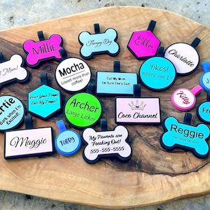 May include: A collection of colorful pet ID tags in various shapes, including bones, circles, and rectangles. Each tag features a name or phrase, such as "Millie," "Mocha," and "Reggie," in a playful font. The tags are set on a wooden surface.