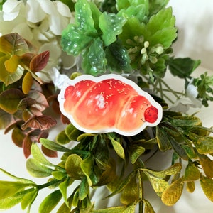 Könnte beinhalten: Ein Aufkleber mit einem Cartoon-Croissant mit einem lächelnden Gesicht. Das Croissant hat eine rot-orange Farbe mit weißen Sesamsamen oben drauf. Der Aufkleber ist von grünen und weißen Kunstpflanzen umgeben.