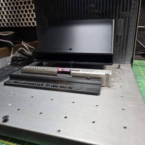 May include: A black rectangular device with a slot is positioned above a metal surface. Below it, a silver and black metal piece is visible, along with a black rectangular piece with text. The setup appears to be for a laser engraving or cutting machine.