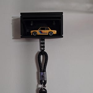 May include: A black key holder with a small yellow car inside. The key holder is attached to a black rope and a black key fob with a silver button.