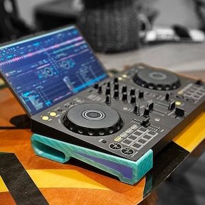 以下が含まれることがあります： 黒いDJコントローラーが、ティールカラーの3Dプリント製スタンドの上に置かれています。コントローラーには2つの円形ジョグホイール、複数のノブ、スライダー、パフォーマンスパッドが搭載されています。コントローラーの後ろにあるノートパソコンの画面には、音楽ミキシングソフトウェアが表示されています。