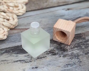 May include: A square, frosted glass bottle with a clear plastic stopper and a separate wooden cube with a hole. The bottle is filled with a light green liquid. A beige woven rope is in the background.